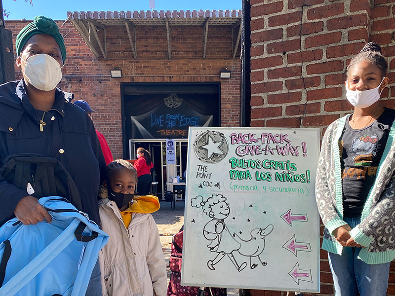 Adult woman holding a backpack with two children next to her at a back pack giveaway.